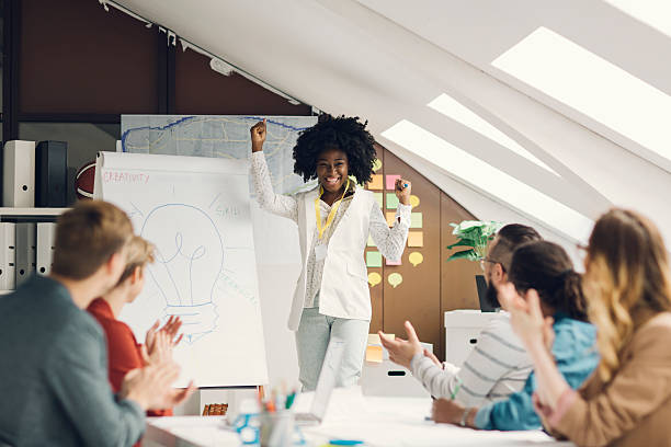 Young african woman has briefing with her coworkers at startup creative agency. He is standing in front of flip chart and talking. Coworkers applauding to her and she is jumping from joy.